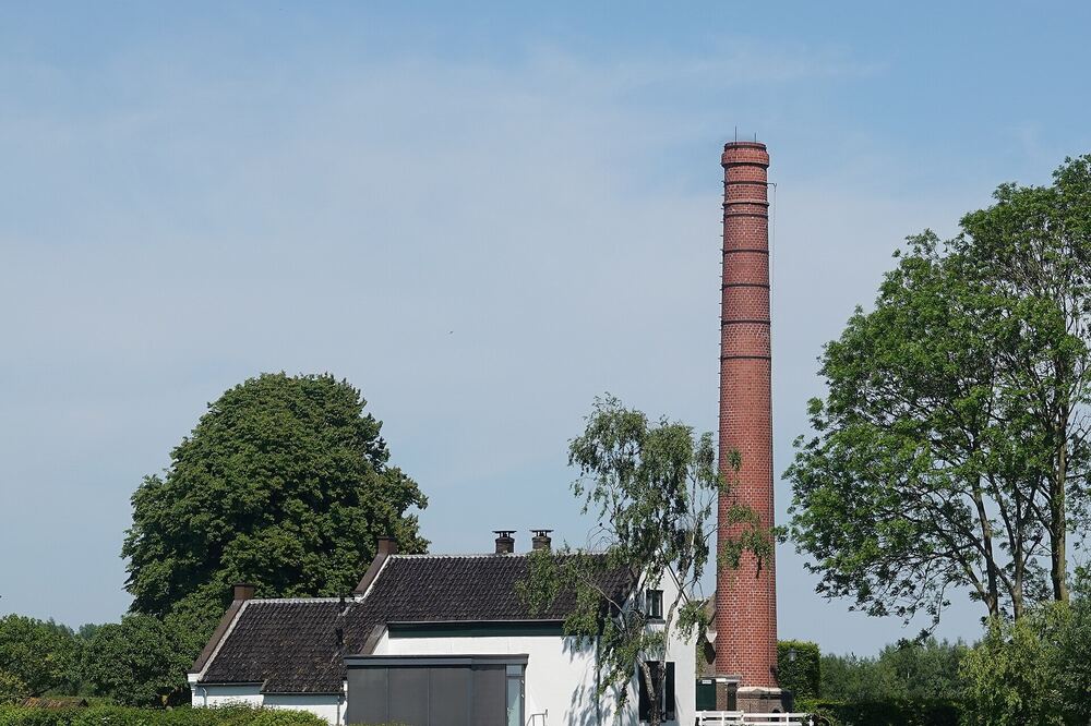 Wit gemaalgebouw met een hoge ronde bakstenen schoorsteen aan de rand van een weiland onder een blauwe lucht bij Putten