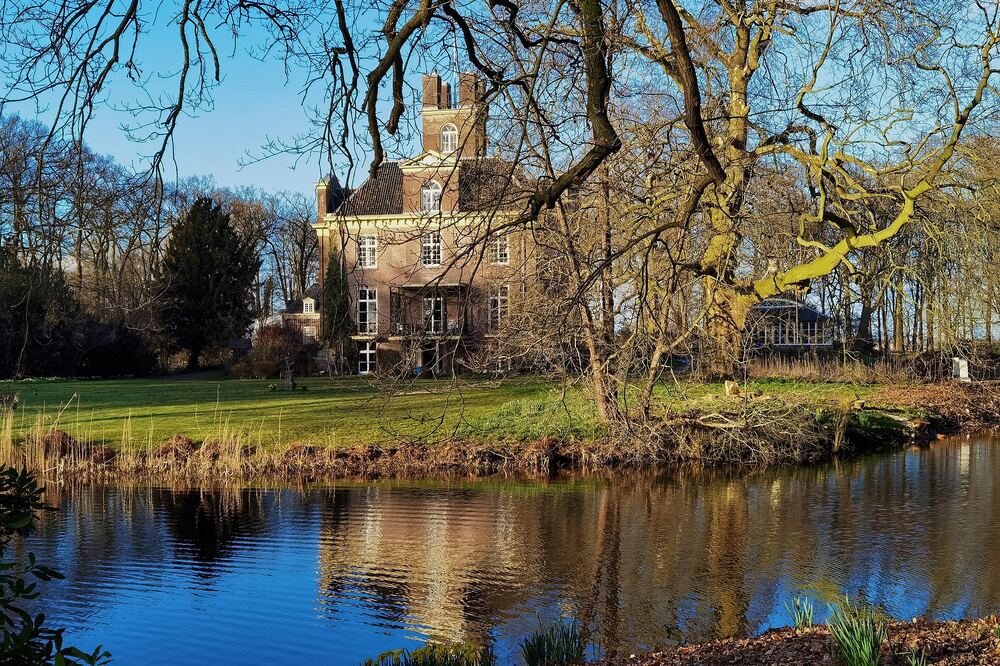 Bakstenen kasteel met toren achter een gazon en een gracht met spiegeling, omlijst door kale boomtakken, op landgoed Oldenaller bij Putten